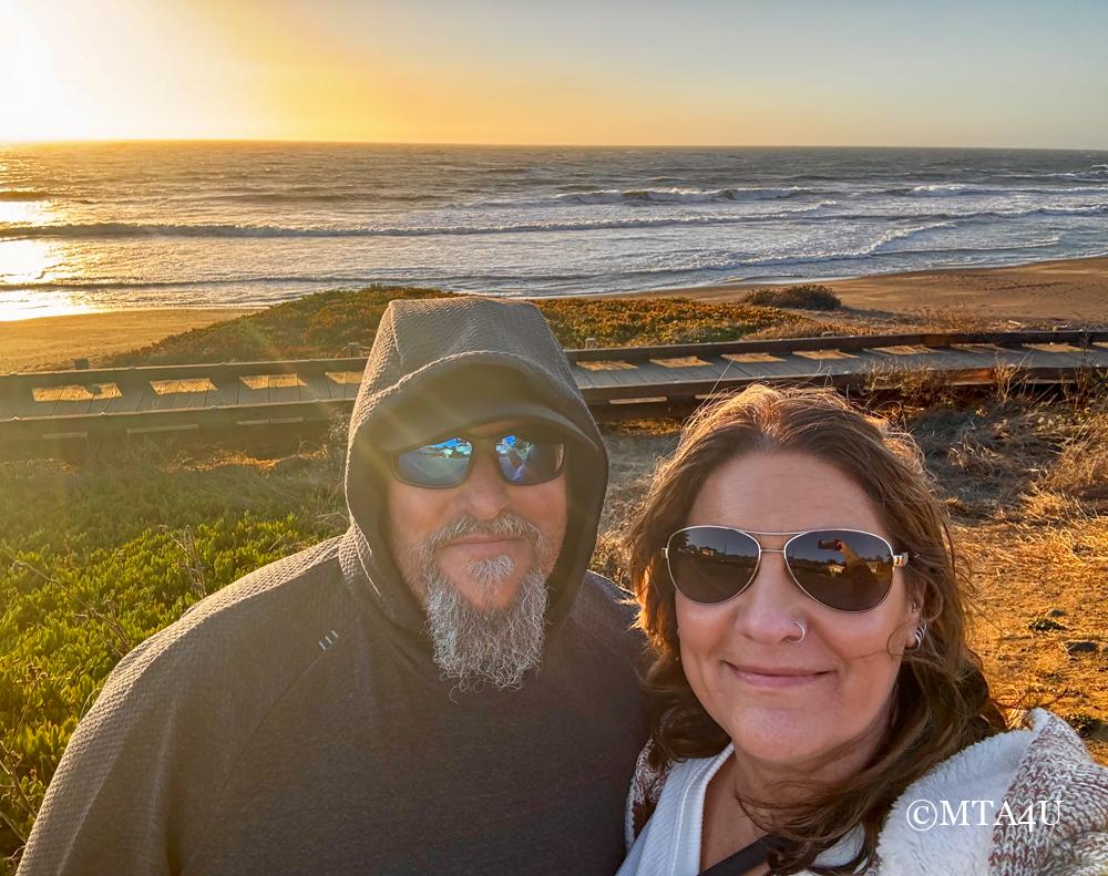 Illya and Noel standing in front of a coastal view at sunset in Cambria, California.