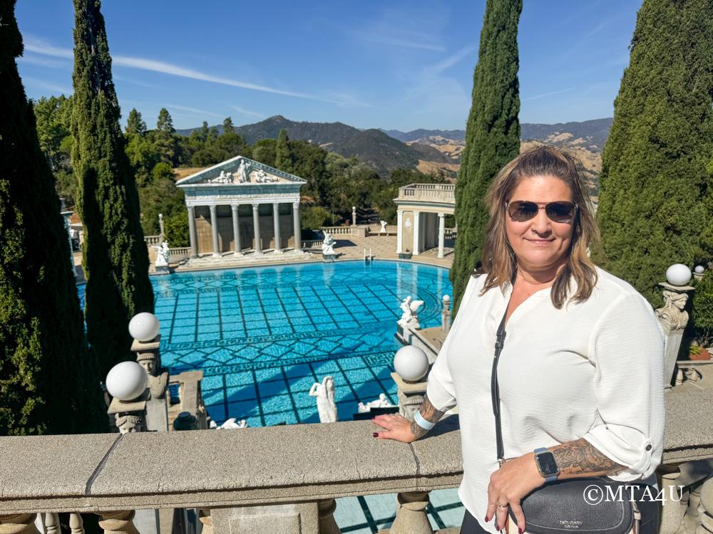 A woman standing in front of the outdoor Neptune Pool at Hearst Castle in San Simeon, California, with the scenic hills in the background.