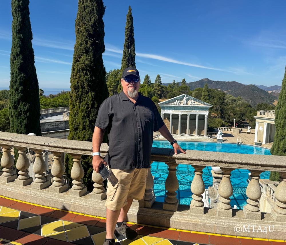 A man standing in front of the outdoor Neptune pool at Hearst Castle in San Simeon, California, with scenic hills in the background.