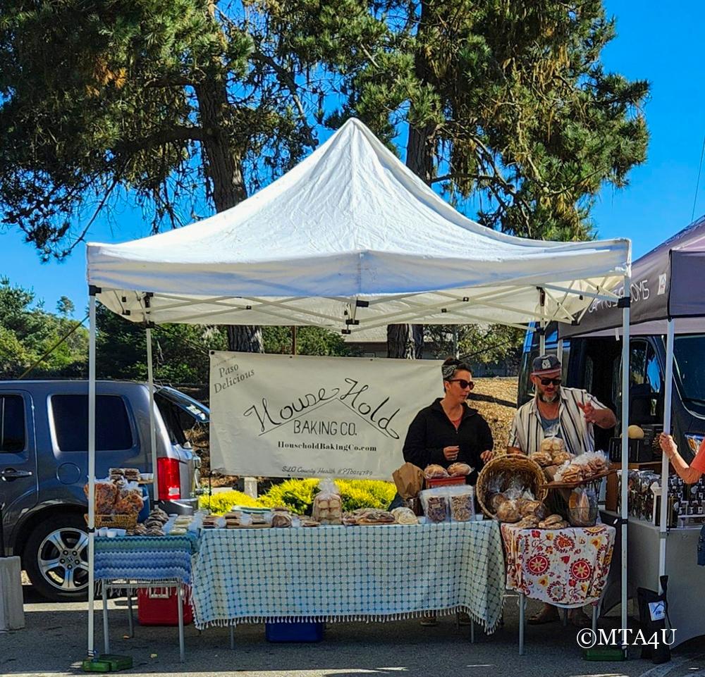 A vendor stall at Cambria Farmers Market with a white canopy and fresh baked goods on display.