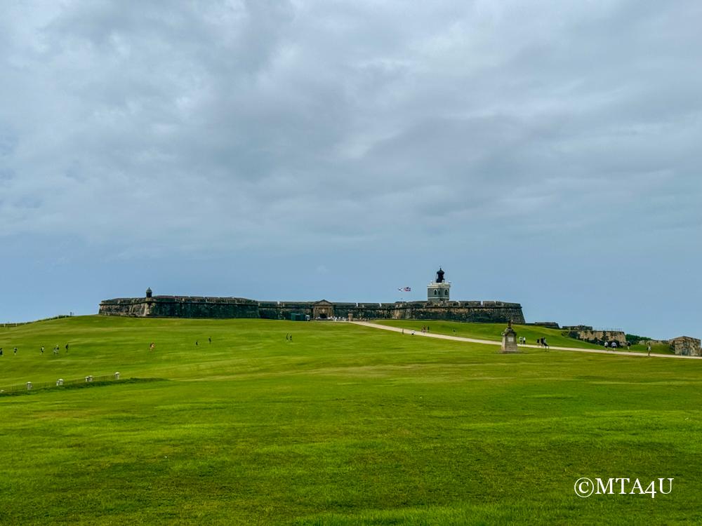 Wide view of El Morro fortress with vast green lawn in the foreground under a cloudy sky in San Juan, Puerto Rico.