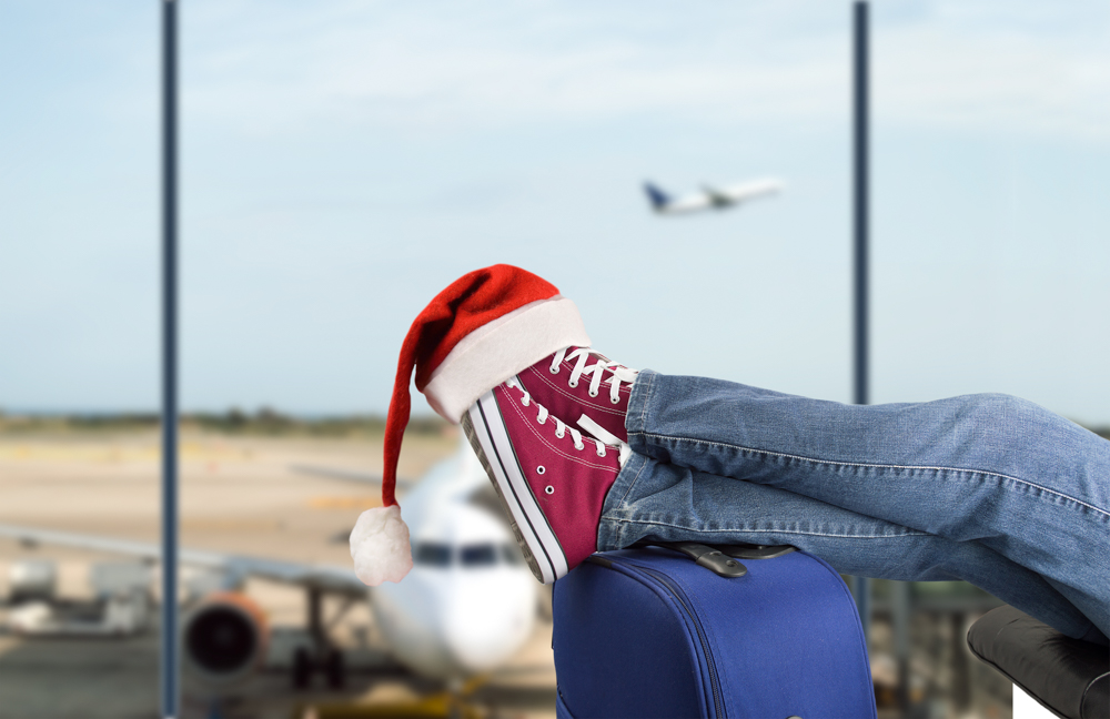 Traveler wearing red sneakers and a Santa hat, resting on a suitcase at an airport terminal with a plane in the background.