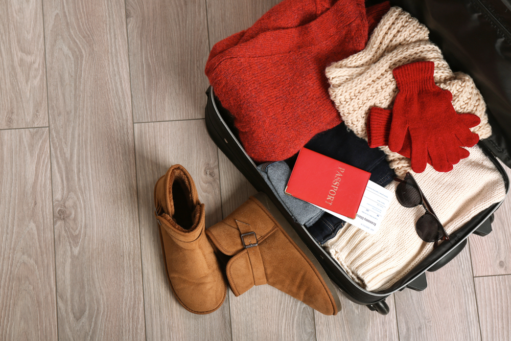 Open suitcase with winter travel essentials, including sweaters, gloves, boots, a passport, and sunglasses.