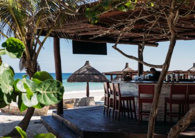 Outdoor beach bar at a Tulum resort with wooden stools, palm trees, and thatched umbrellas.