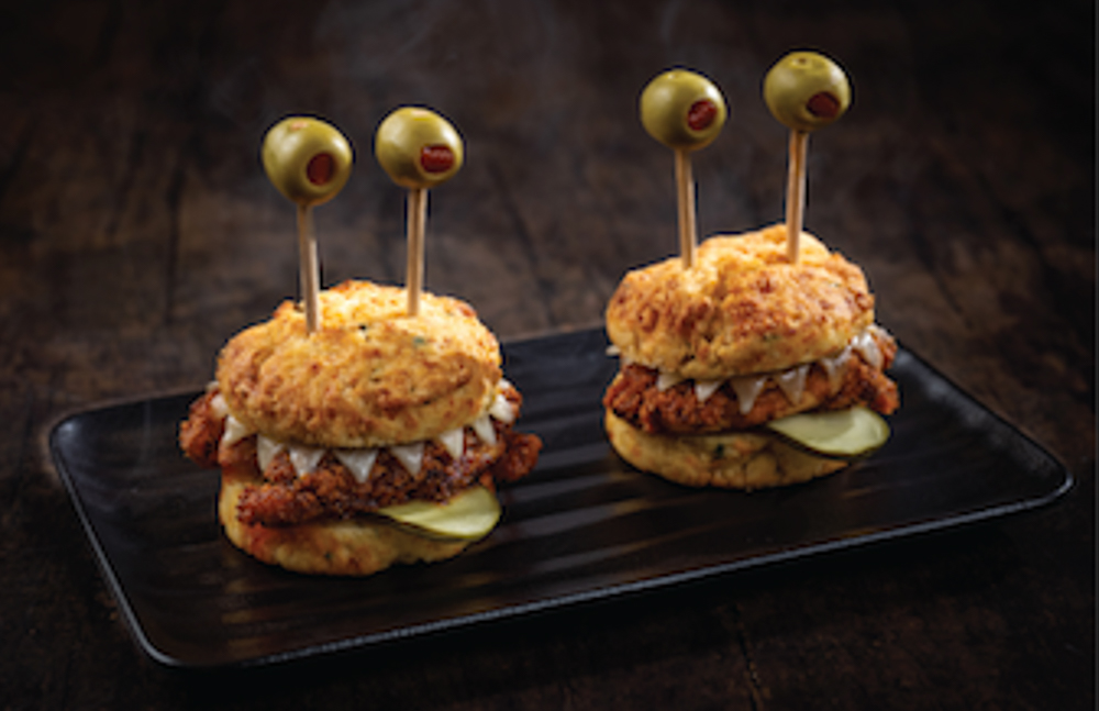 Two Halloween-themed chicken sliders styled with olive "eyeballs" on top, served on a black plate with pickles and biscuit buns.