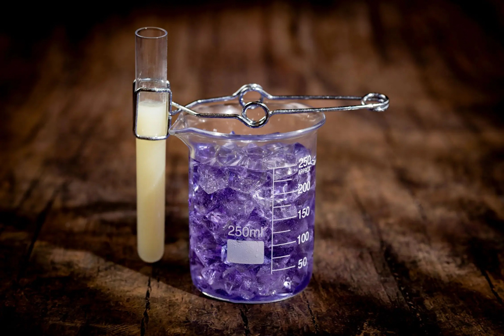 A spooky purple cocktail served in a 250ml beaker filled with ice, with a glowing yellow test tube attached using lab-style metal tongs.