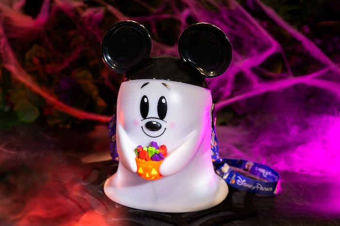 A glowing Mickey-shaped ghost popcorn bucket holding a candy-filled pumpkin, featuring Mickey ears and a blue Disney Parks lanyard, set against a spooky background with purple fog and spider webs.