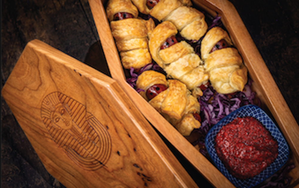 A wooden sarcophagus box filled with puff pastry-wrapped mummy-style hot dogs resting on shredded purple cabbage, served with a blood-red dipping sauce in a small blue dish.<br>
