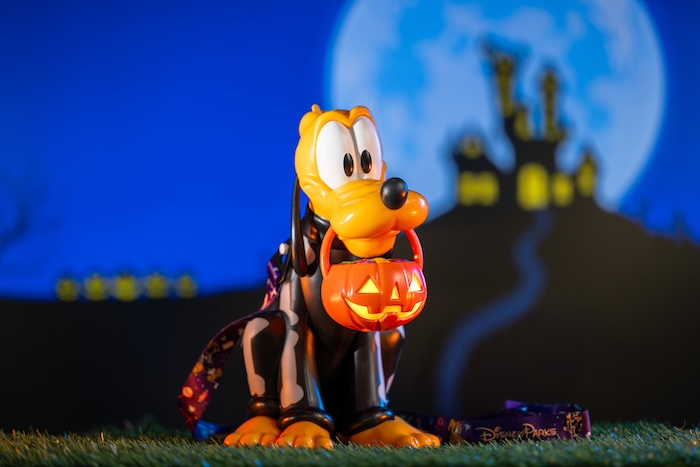 Pluto dressed in a Halloween costume holding a glowing jack-o’-lantern bucket in his mouth, with a spooky blue-lit castle silhouette in the background.