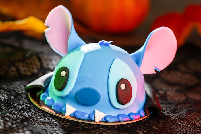 A dome-shaped cake decorated as Vampire Stitch with large ears, fangs, and a cape, styled on a black lace tablecloth with autumn decor in the background.